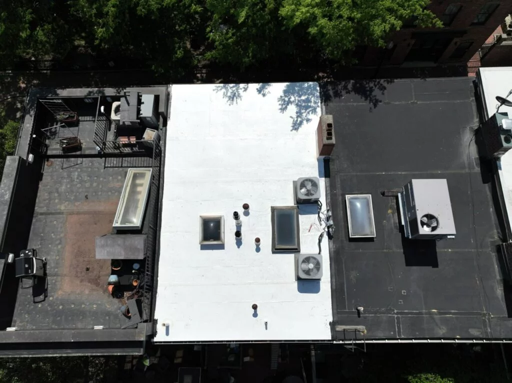 Aerial view of a white flat roof with skylights, vents, and HVAC units, contrasting with adjacent black roofs