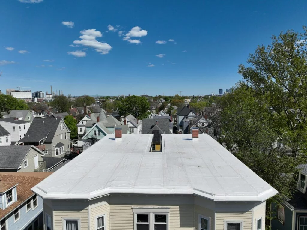 Flat white roof of a residential building with chimneys, overlooking a neighborhood with a city skyline in the distance